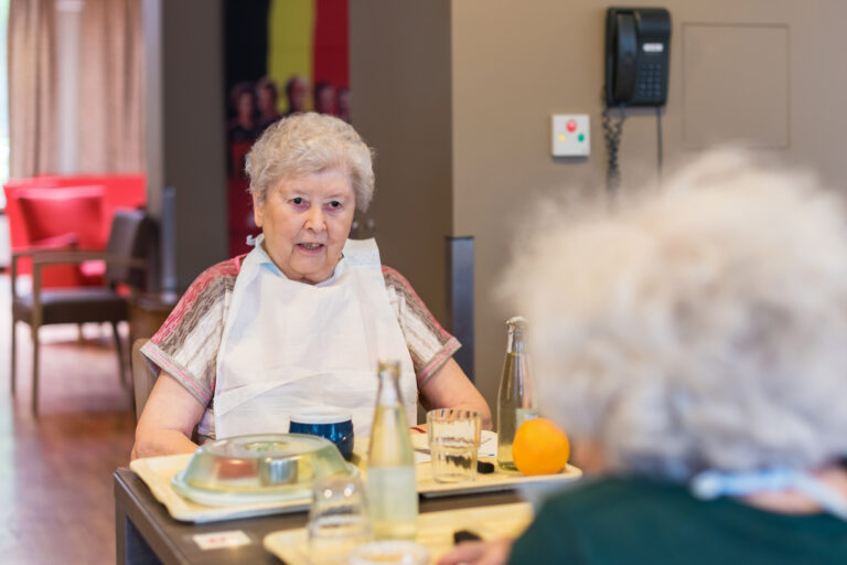 Une dame âgée de face et une dame de dos entrain de manger dans le réfectoire avec des plateaux repas